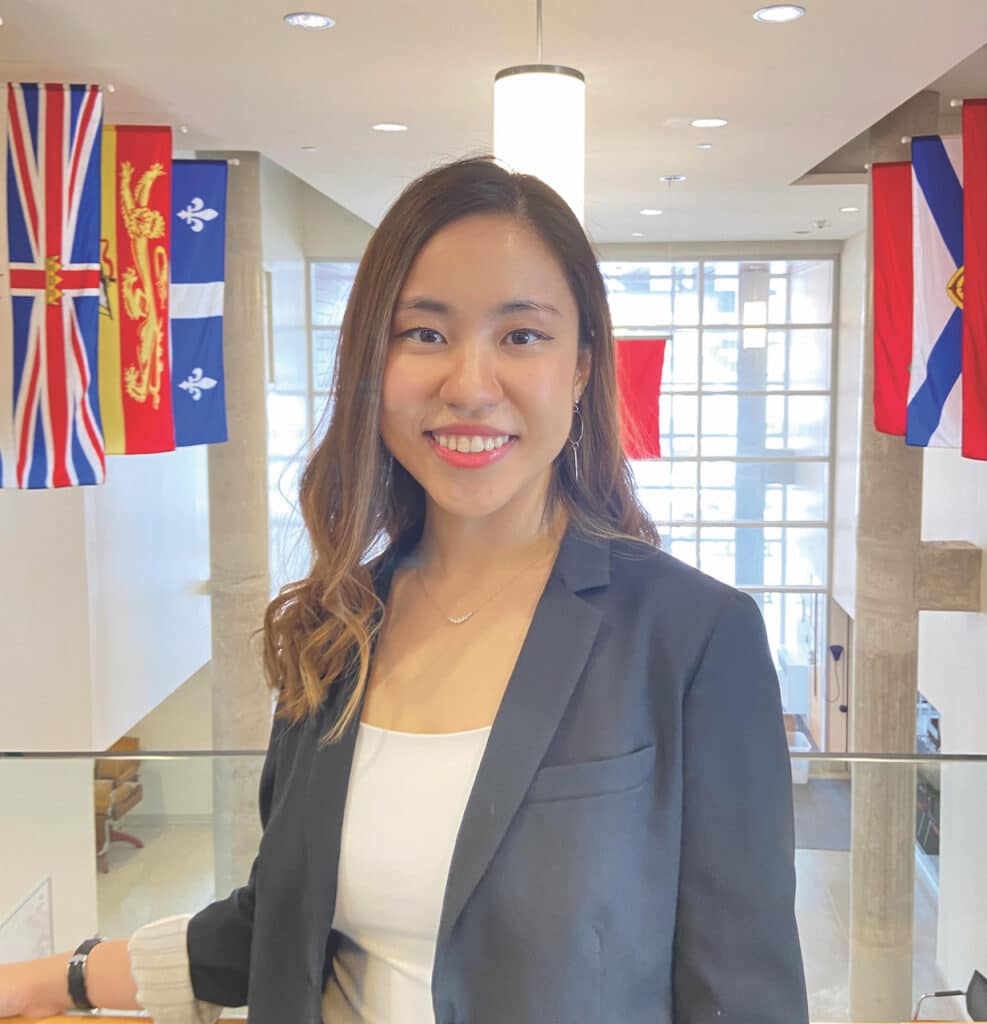 Photograph of the author, Jenny Lee, smiling in a large indoor space with provincial flags behind her.