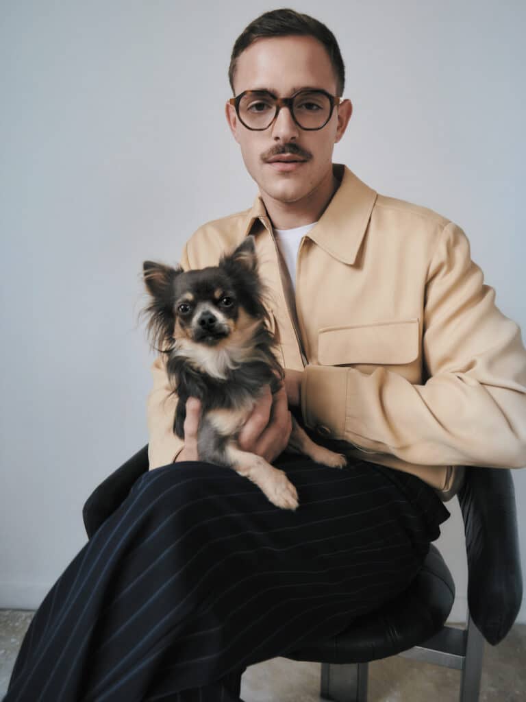 Stylish man in a beige jacket holding a small dog, wearing GIGI STUDIOS tortoiseshell glasses