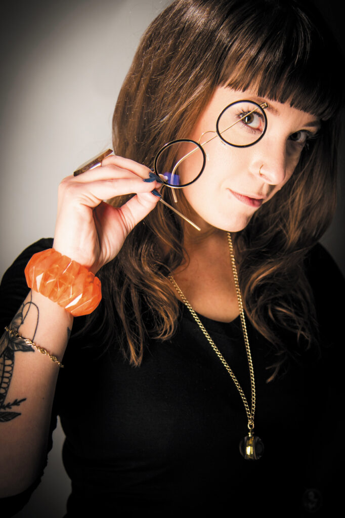 Artistic photograph of Sarah Bureau, holding a pair of round glasses framing one eye.
