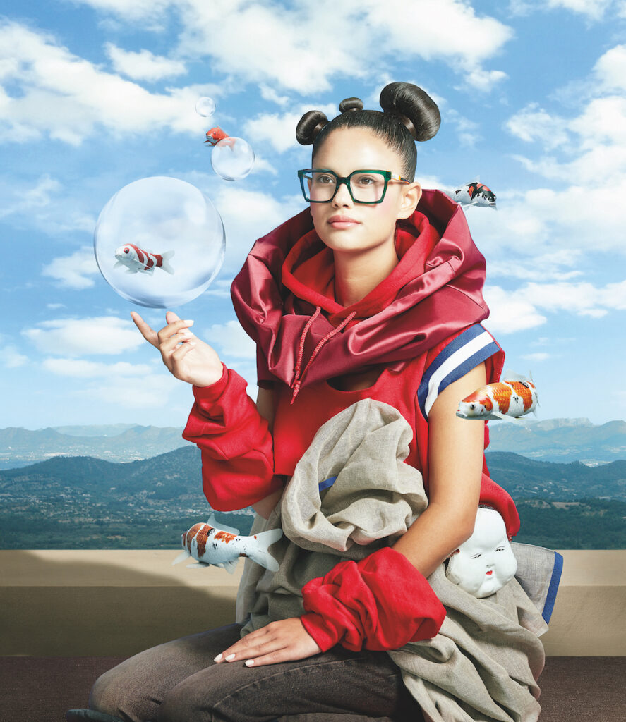 Mixed-media image showing a model wearing bold Spanish eyewear, surrounded by floating bubbles and fish, against a village hillscape.