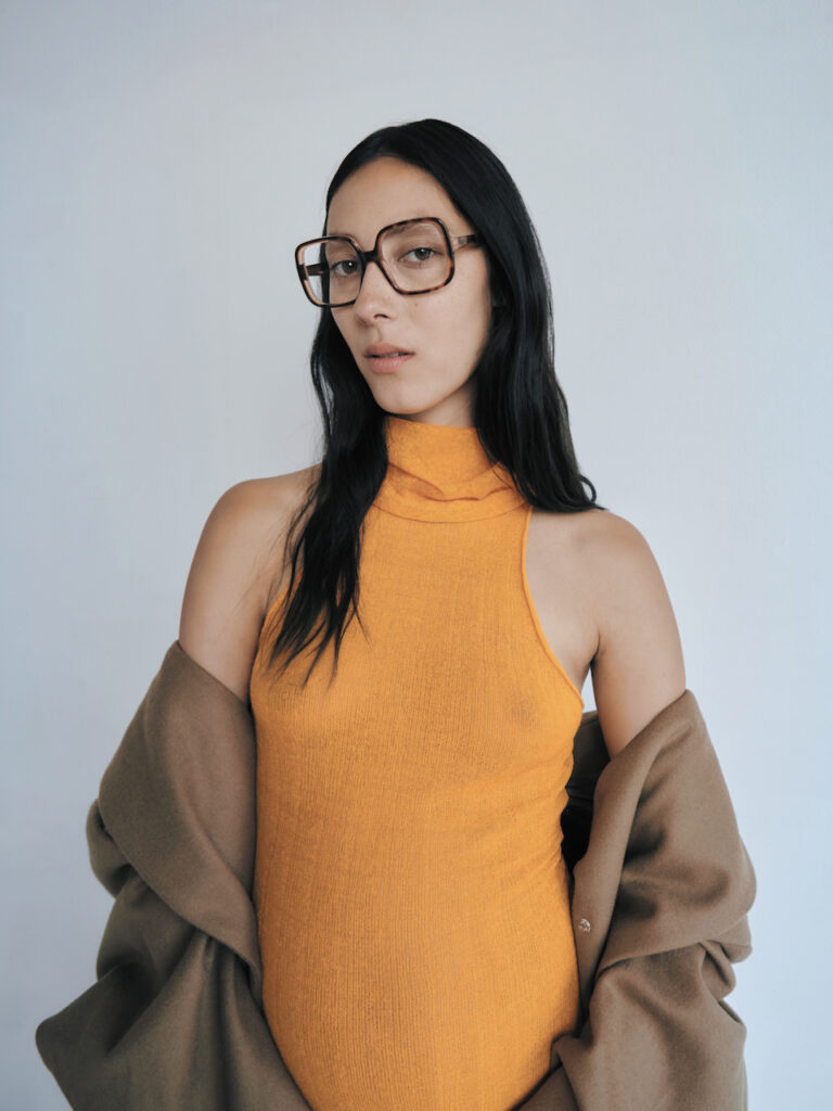 Trendy woman in an orange turtleneck and oversized GIGI STUDIOS glasses posing in a studio.